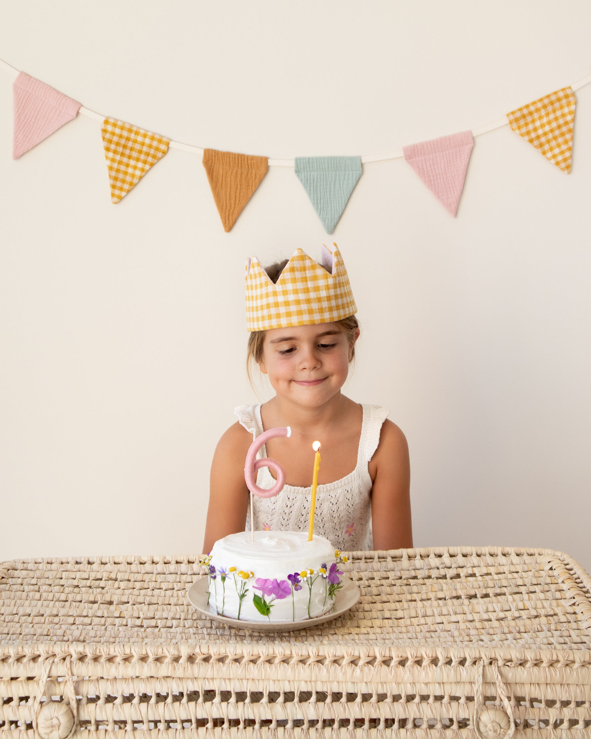 Coroa de aniversário reversível em musselina vichy mostarda, bolo florido com um topper número 6 e uma vela de cera de abelha amarela, grinalda com bandeirolas coloridas em musselina