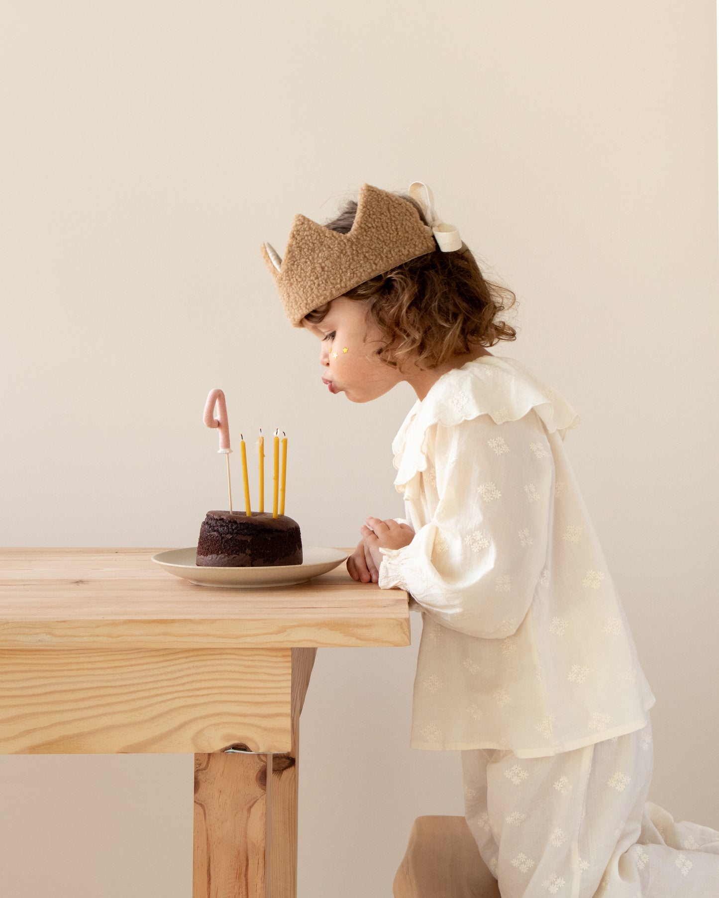 coroa de aniversário reversível em pelo carneirinho castanho, menina a soprar velas num bolo de chocolate com topper número 4 rosa